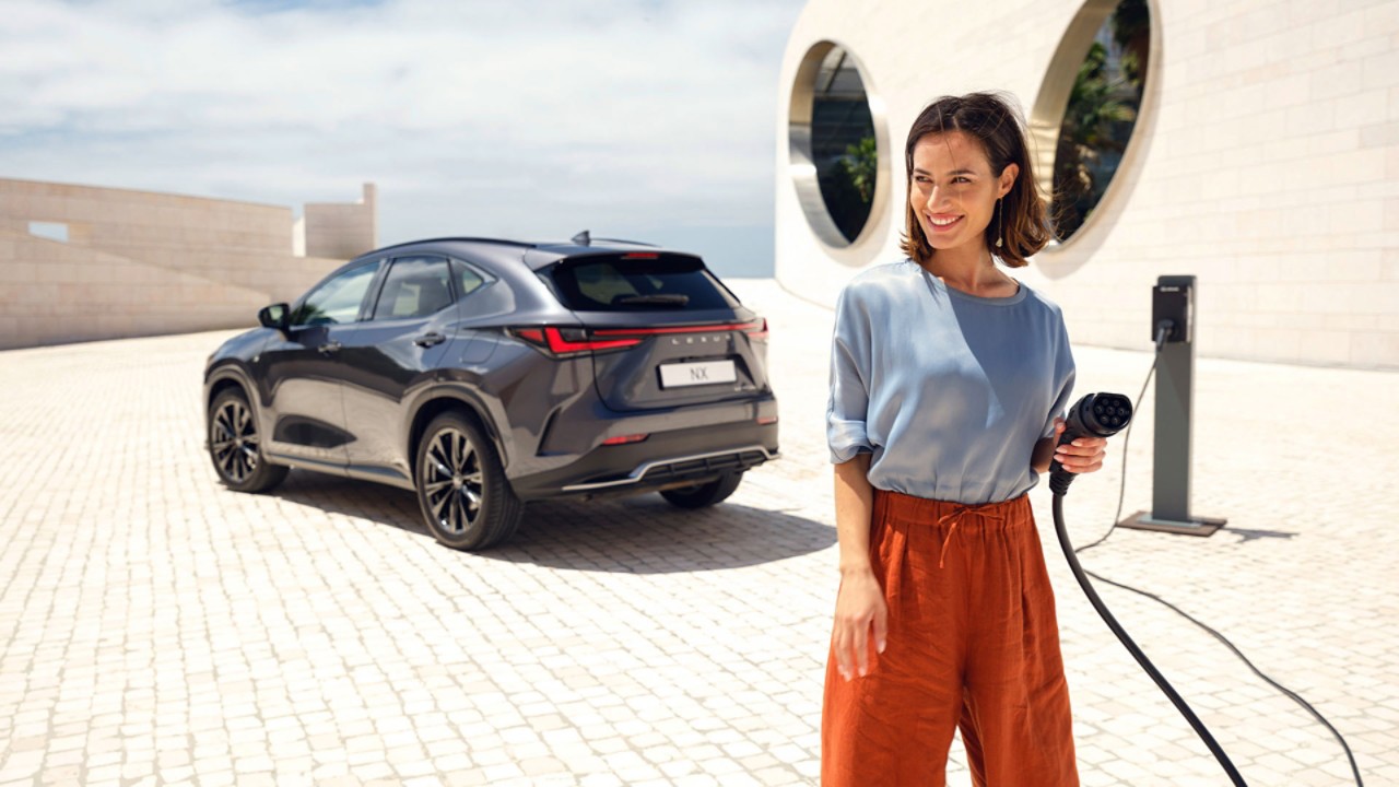 A person holding a charging plug in the foreground of a parked Lexus NX